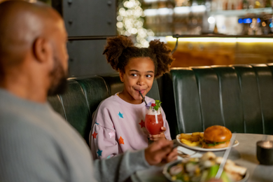 A child slurping a drink and having a meal with her father