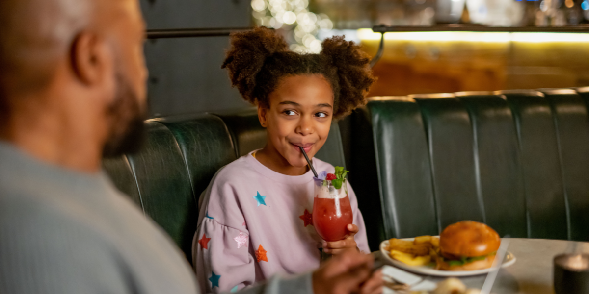 A child slurping a drink and having a meal with her father